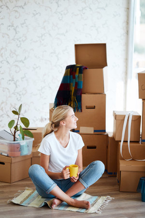 smiling woman with tea in hand sitting on floor of new apartment, pile of moving boxes on background