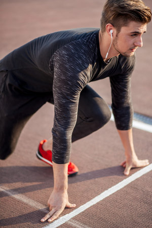 young man wearing black clothes and red shoes in starting position for running on sports track in stadiumの写真素材
