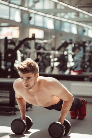 Strong, handsome fitness instructor doing push-ups on dumbbells in a gym as bodybuilding exercise,...