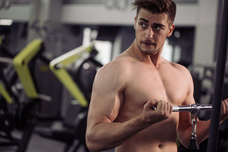 Handsome young guy pumping up upper body muscles doing exercises on gym mashine with naked torso,...