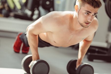 Strong, handsome fitness instructor doing push-ups on dumbbells in a gym as bodybuilding exercise,...