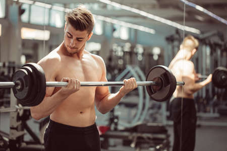 Shortless man in black sportive pants with naked muscular torso holds a barbell on gym equipment background.の写真素材