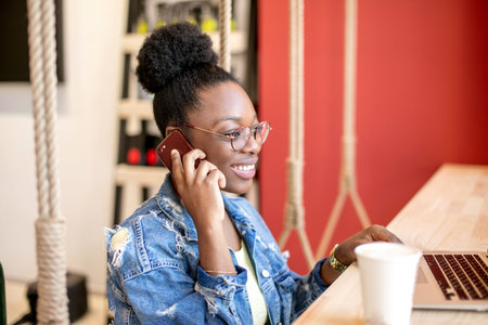 Hipster dark-skinned woman with Afro hairstyle talking on smart phone at cafe. Young woman sitting...