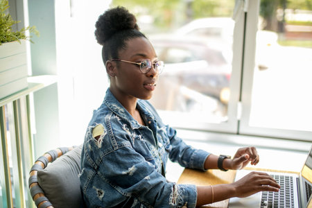 Side view of African woman in denim jacket sitting near the window and using laptop in cafe....