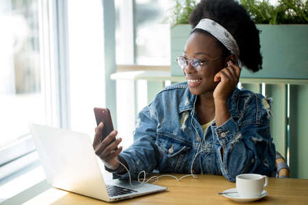 Young african customer client female in spectacles and jeans outfit beaming with joy, spending free time shopping online while sitting in cafe.の写真素材