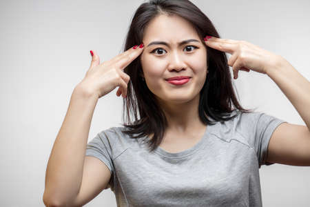 Discontent frowning young Asian woman wearing grey t-shirt, looking at camera, standing and making suicide gesture. Stress and problem conceptの写真素材