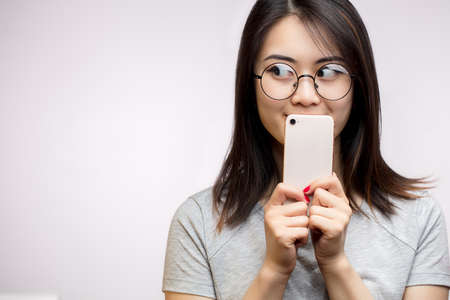 Flirting asian woman with tricky eyes hiding mouth with smart phone isolated over white background in studioの写真素材