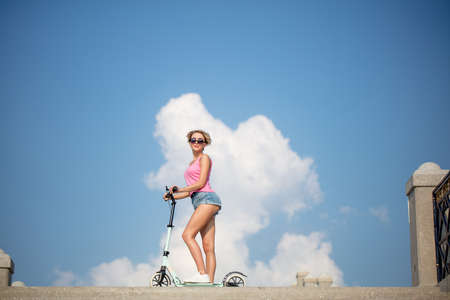Joyful blonde woman in pink top, sunglesses and jeans shorts riding a kick scooter on sky backgroundの写真素材