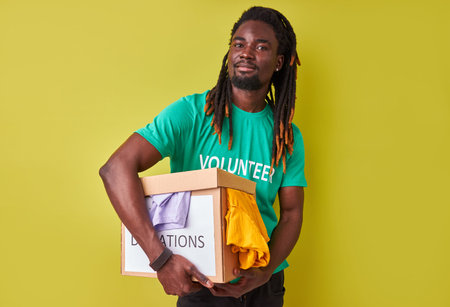 portrait of black afro volunteer man with box full of clothes, young male with dreadlocks engaged in...