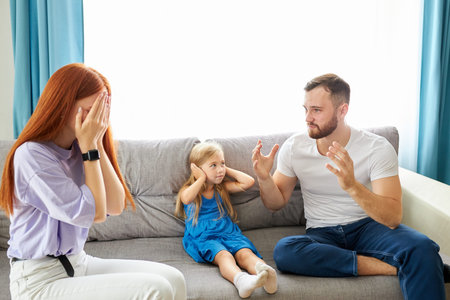 sad, desperate little girl during parents quarrel, she clog the ears sitting on sofa at home, angry parents fighting. worried upset small daughter hurt by fathers and mothers break up or divorceの写真素材