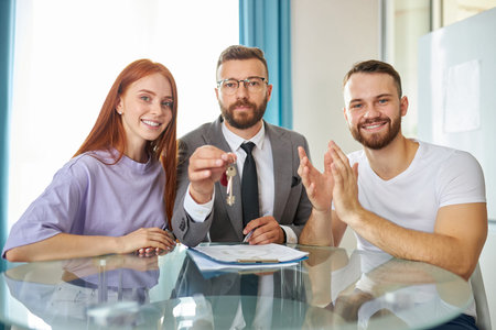 portrait of happy young newly married family with real estate agent sitting at table together, agent in formal suit hold keys in hands, they smile at cameraの写真素材