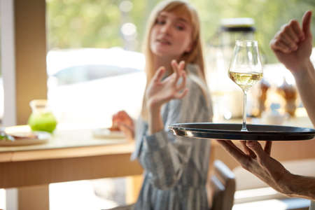 glad waiter bring glass of champagne on tray to customers, treats women in restaurant. caucasian woman is thankful for service. focus on glass with beverageの写真素材