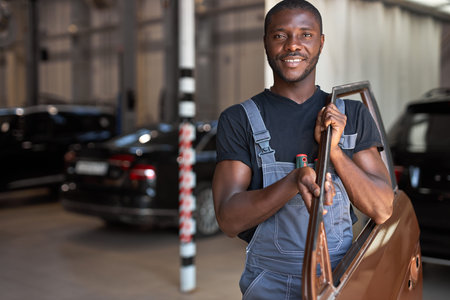 young african auto mechanic at work with separate part of the machine, posing at camera, going to...