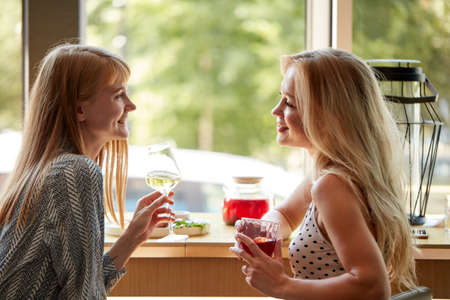 side view on happy blonde women talking and drinking champagne in cafe, at meeting after work. lunch time. relaxed females share news, smiling friends, lifestyle conceptの写真素材