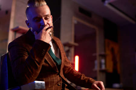 confident wealthy caucasian male with beard in formal stylish suit sit smoking cigar, relax. isolated in dark smoky roomの写真素材