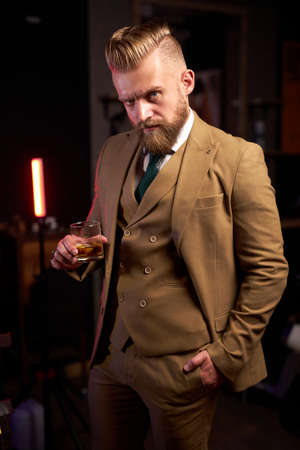 young caucasian bearded business man drinking glass of whiskey indoors in dark room, wearing formal suitの写真素材