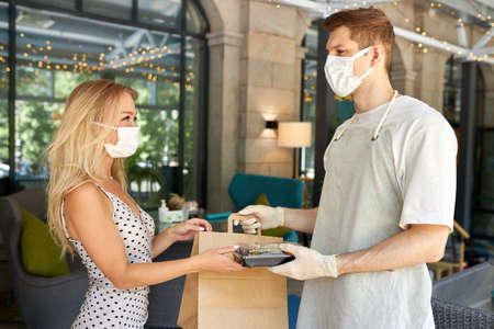 adorable woman customer take her order in restaurant during coronavirus, waiter staff of cafe give food in container wearing protective gloves, in medical maskの写真素材
