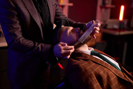 hairstyling process. barber cutting beard of caucasian male with the use of scissors, client sits in an armchairの写真素材