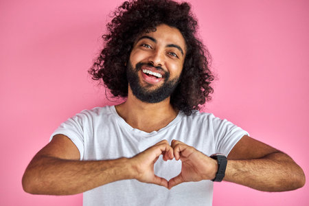 young indian or arabic man smiling in love doing heart symbol shape with hands, happy curly bearded male isolated on pink background. romantic conceptの写真素材