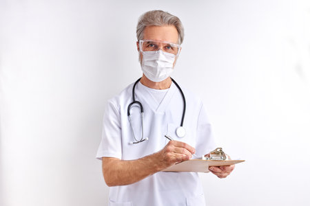 elderly doctor man makes notes in tablet wearing stethoscope and medical mask isolated on white studio background, medicine and healthcare conceptの写真素材