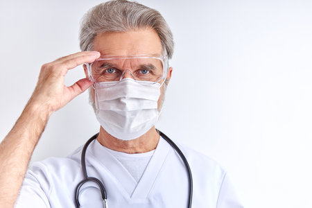 caucasian senior doctor male in protective eyeglasses goggles, wearing it before work with patients, looks at cameraの写真素材