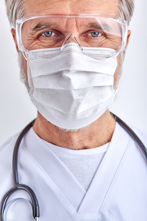 confident male doctor in mask, mature elderly male looks at camera wearing white medical suit and goggles, using stethoscope, white backgroundの写真素材