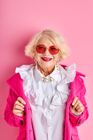 happy optimistic aged lady in blouse and coat smiles at camera, wearing sunglasses, isolated over pink backgroundの写真素材
