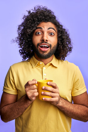emotional arabic male with mug in hands, isolated over purple background. handsome guy with curly hair look at camera with opened eyes, surprisedの写真素材