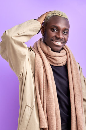 portrait of handsome afro american male in stylish wear posing at camera isolated on purple studio background, young male has perfect toothy smile, wearing beige scarf and coatの写真素材