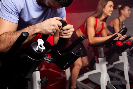 close up hands of man biking in spinning class, group of people exercising on stationary bike in the background at gymの写真素材