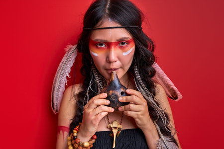 shaman woman using authentic indian whistle isolated on red background, female with feathers on head...