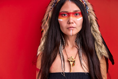 wild female with colourful make-up isolated in studio on red background, youngwoman is wearing hat made from feathers on head, looks at cameraの写真素材
