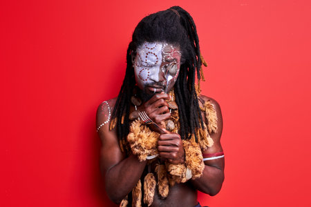 african native guy trying to use modern cutlery, holding spoon and fork in hands, isolated over red...