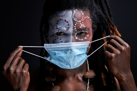 primitive afro american man is wearing protective medical mask on painted face, isolated over black studio background. naked male with dreadlocks keep safe and healthyの写真素材