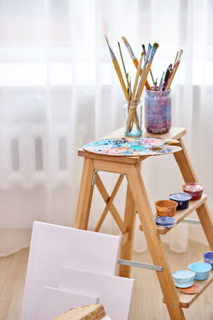 close-up photo of paints, paintbrushes, easel and canvas in light art studio roomの写真素材