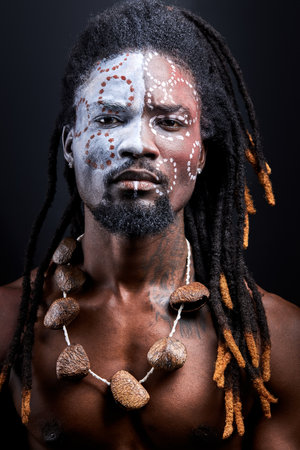 african shirtless guy with colourful make-up isolated on dark studio background, young male tribal has ethnic traditional chainの写真素材