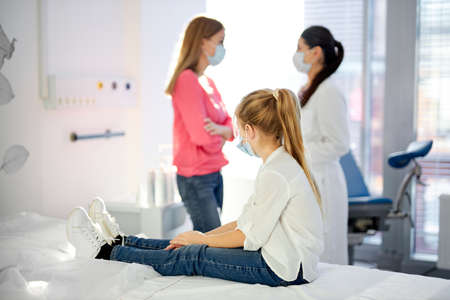 sick girl sits looking at mother talking with doctor in hospital, waiting for solution, how to treat, in medical masks. coronavirus, covid-19. focus on girlの写真素材