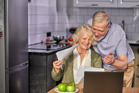 elderly man and woman managing family monthly budget together, positive married couple using computer banking application, counting bills in the kitchen discussingの写真素材