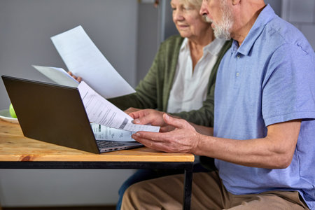cropped elderly man and woman managing family monthly budget together, focused married couple using computer banking application, counting bills in the kitchen. focus on laptop and handsの写真素材