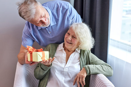 senior female person with satisfied face getting present box by her nic handsome husband,...