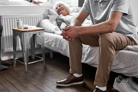 depressed man sits near his sick elderly wife lying on bed suffering from disease. in hospitalの写真素材