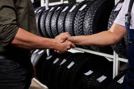 cropped customer and salesman shaking hands against the background of car tires. make purchase after choosing the best oneの写真素材