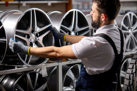 side view on bearded male auto mechanic repairman examining car rims, checking the assortment in his shop, at work place, wearing uniformの写真素材