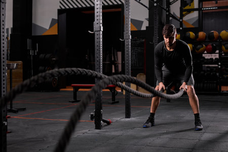 strong young man working out with battle ropes at cross fit gym. muscular caucasian sportsman doing...