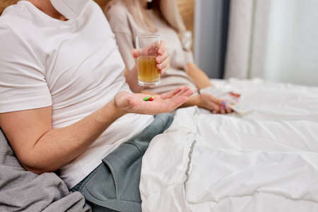 cropped couple at home with seasonal winter cold illness disease lie on bed taking medications, support and help each other, at home. coronavirus, covid-19, sickness conceptの写真素材