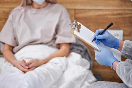 doctor asks female patient about state of health, professional doctor taking notes, covid-19. stay at home concept during Coronavirus pandemic and self isolation. focus on pen and paper, handsの写真素材