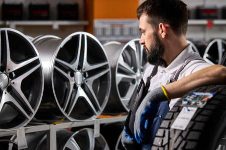 adult auto mechanic man in uniform have rest after hard working day, standing by rack of car wheels. auto, car, transport conceptの写真素材