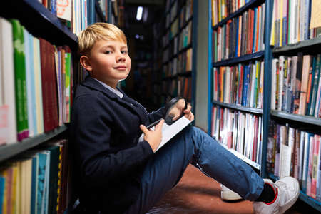 boy carefully reads every word in book using a magnifying glass, while sitting on the floor between the shelves in libraryの写真素材