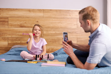 modern daddy take photo of his trendy daughter using cosmetics, man and child sit on bed together,...