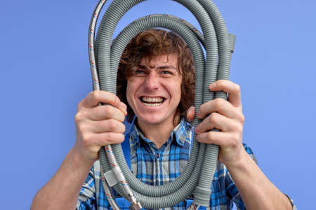 young plumber man holding hose and looking at camera through it, strong confident sanitary engineer irritated, doesnt know reason of leakage or accidentの写真素材